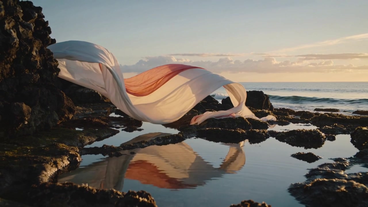 Onshore breeze lifting flowing fabric into loops across rocky tidal shore, reflecting in tide pool