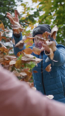 primer plano de parientes disfrutando de un momento lúdico al aire libre con el follaje seco de otoño, los miembros de la familia lanzan hojas alegremente el uno al otro, rodeados de árboles verdes y dorados