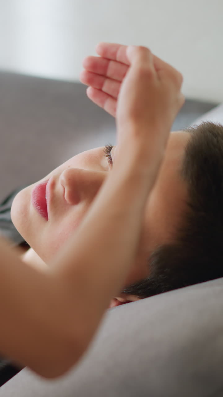 Close-up of someone placing hand on forehead of sick child lying on the couch, the child looks weak, and someone is observing with concern, offering care