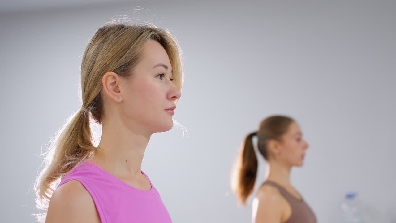 러시아 여성들, 한 명은 분홍색, 다른 한 명은 갈색 스포츠 옷을 입고, 체육관에서 일치하여 피트니스 루틴을 연습합니다.