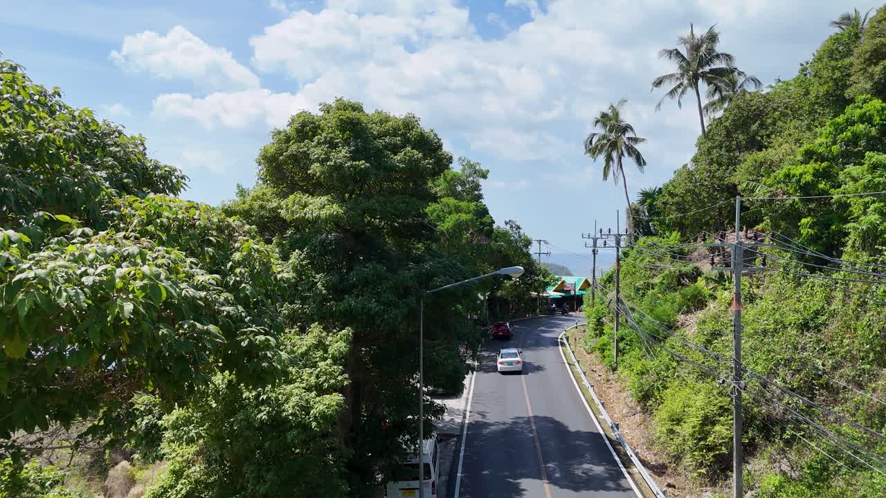 푸켓의 나무로 둘러싸인 도로를 가로질러 달리는 자동차는 밝은 빛 아래 활기찬 초록색과 조용한 열대 분위기를 보여줍니다.