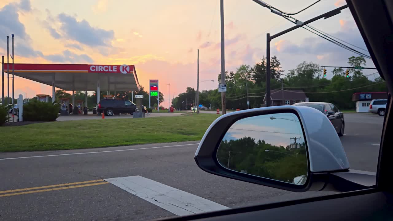 Stock video of a white SUV turning onto a road at sunset near a Circle K gas station. Captures a calm suburban moment with vehicles, power lines, and a colorful sky in the background.