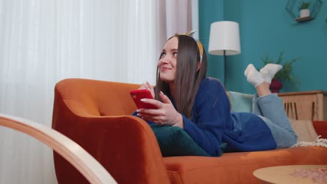 joyful young adult woman lying on sofa using smartphone watching video online shopping tapping