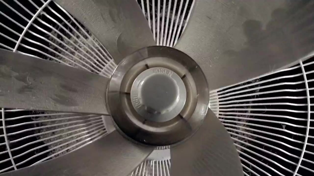 Dusty oscillating fan blades turn behind a metal grille in close up.