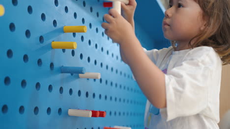 little girl stick wooden multicolor sticks into peg board wall with holes at inside playroom - a handheld close-up shot