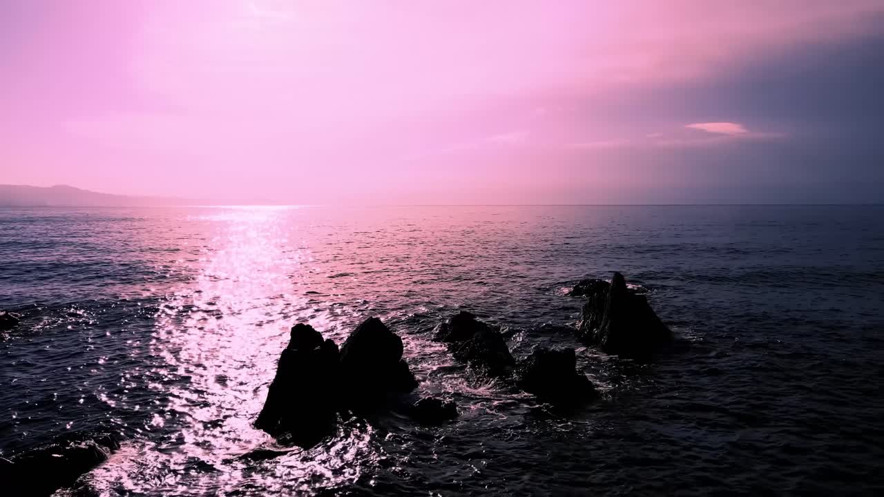 Drone video captures Italy's ocean at sunset with spectacular pink hues reflecting on the water, highlighting rugged rocks under a tranquil sky, creating a serene atmosphere