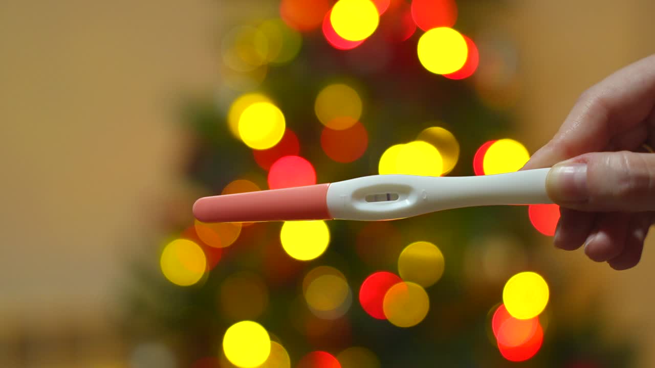 Female hand holds a pregnancy test with two lines which means is positive in front of a Christmas tree changing the lights in a bokeh effect
