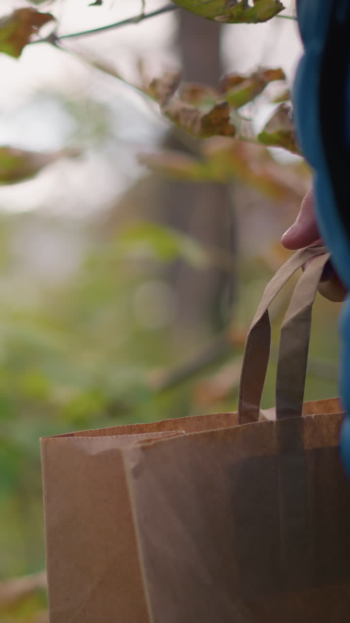 파란색 재을 입은 사람이 는 동안 종이 봉지를 들고, 근처의 나무에서 잎을 잘라서 봉지에 넣는 부분적인 근접 시각, 배경에 흐릿한 활기찬 초록색