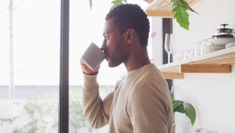 Happy-african-american-man-standing-in-kitchen,-drinking-coffee