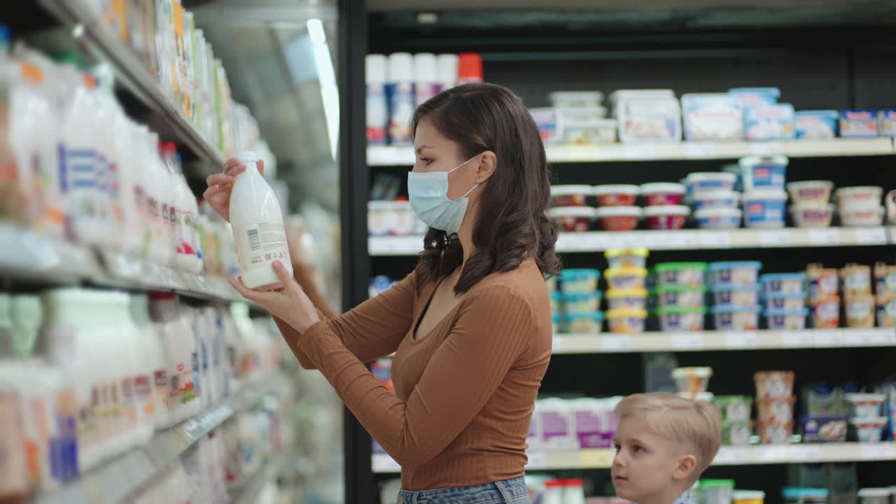 una mujer con una máscara en un supermercado elige una bebida cerca del refrigerador de leche o yogur
