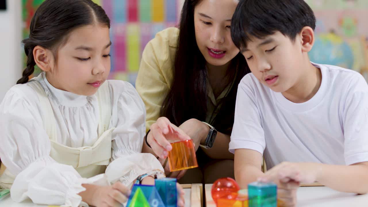 アジア系の教師が、明るい教室でカラフルな幾何学的な図形を使って2人の小学生を指導しています。