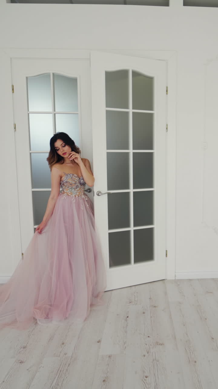 Beautiful bride in wedding dress is standing near the door in the light room. Gorgeous model in luxury dress is posing in studio. Vertical video