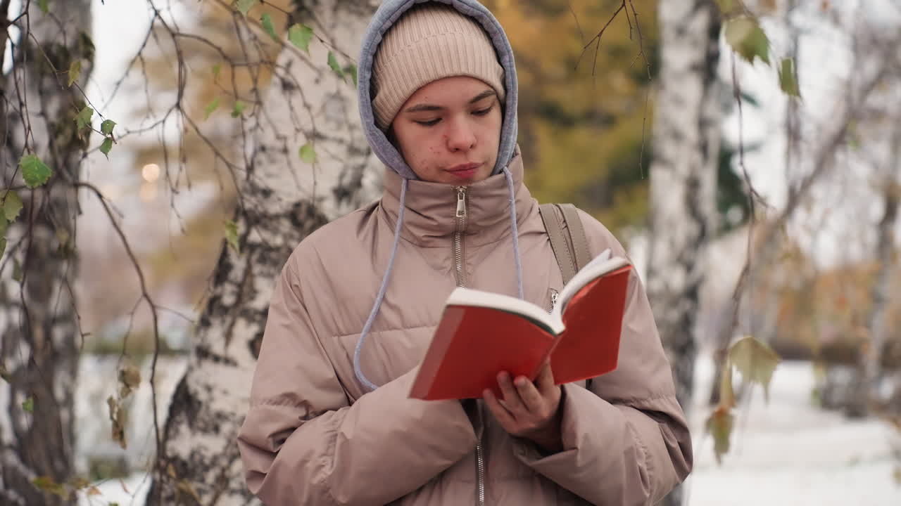 Light skin lady in warm jacket and beanie flipping through red book while standing beside tree on snow-dusted ground, surrounded by autumn foliage and soft background of birch trunks and branches