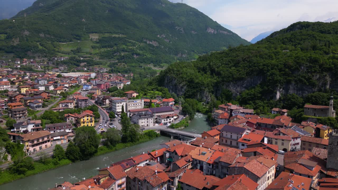 Expansive drone shot of village beside a mountain ridge, with curved river and urban spread. Tilt down drone descend. Shot in Cividate Camuno, Italy (Cividate Camuno, Italia)