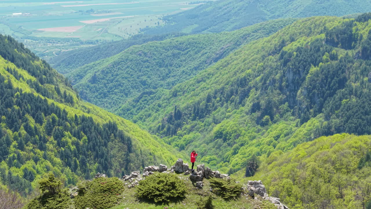 Drone view of a woman at a viewpoint in the Balkan Mountains, Bulgaria. The landscape features meadows, forests, and winding paths under a soft spring sky.
