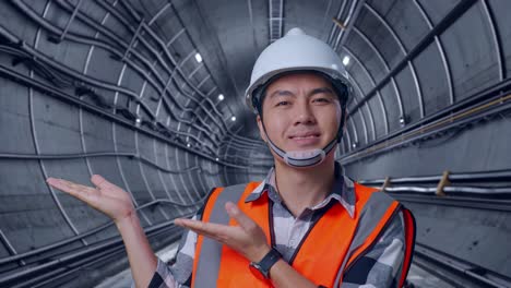 close up of asian male engineer with safety helmet smiling and pointing to side while standing in underground subway tunnel