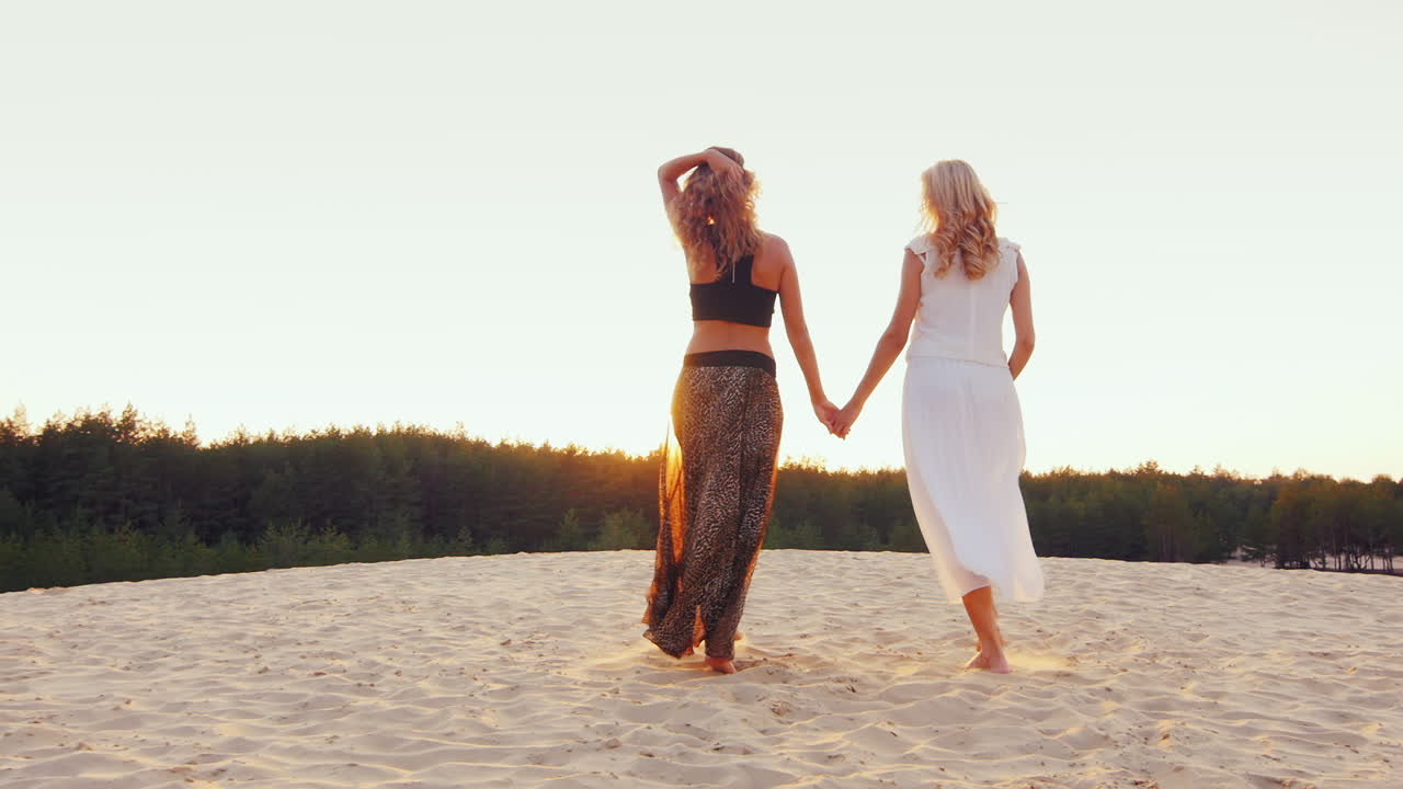 아름다운 드레스를 입은 두 명의 날씬한 여성이 모래사장을 지나 일몰의 뒷모습으로 들어갑니다
