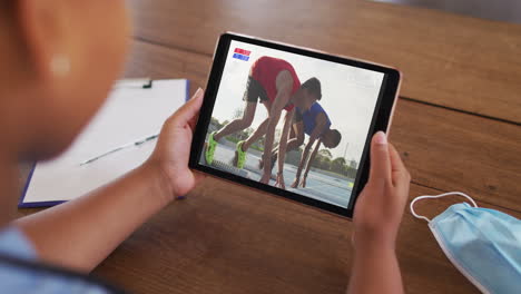 compuesto de una mujer sentada en una mesa con una máscara facial, viendo un evento de atletismo en la tableta