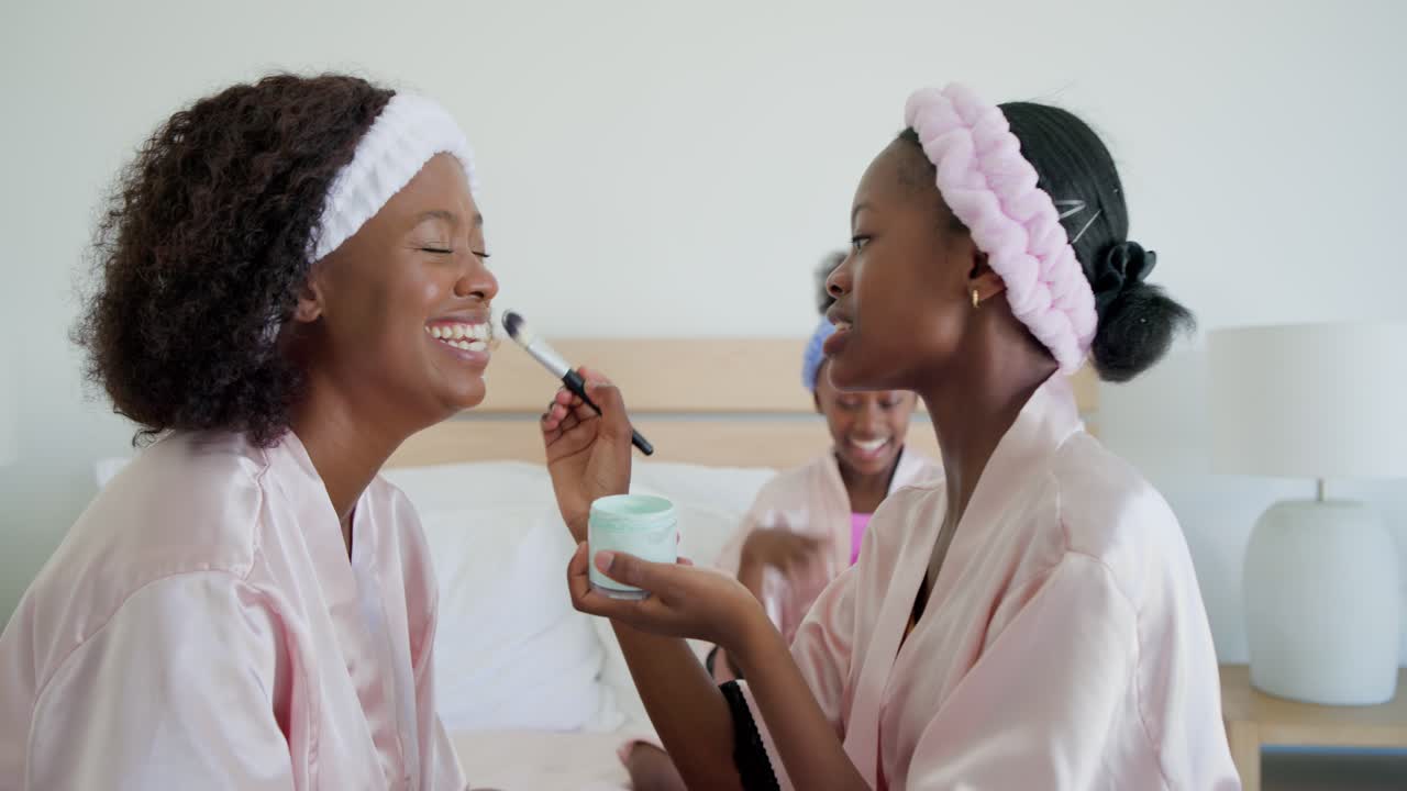 Teenage daughter in pink robes on bed dipping brush into jar, pampering mother while child watching