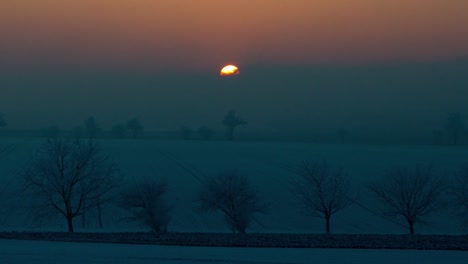 El-Tiempo-De-Puesta-De-Sol-Cae-En-Un-Paisaje-Nevado-Con-Campos-Y-árboles,-La-Cámara-Se-Acerca-Y-Sigue-El-Sol