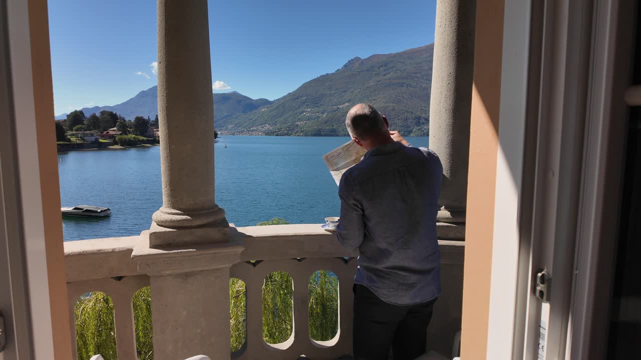 A pan American shot of a man drinking coffee on a balcony overlooking Lake Como, planning a trip using a map