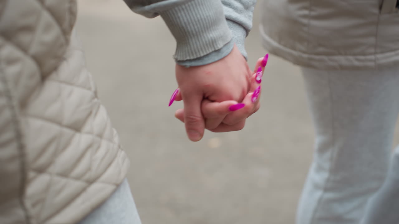 Couple walking hand in hand along tarred road with woman's nails painted purple, fingers intertwined, captured in close-up with dry foliage blurred on ground