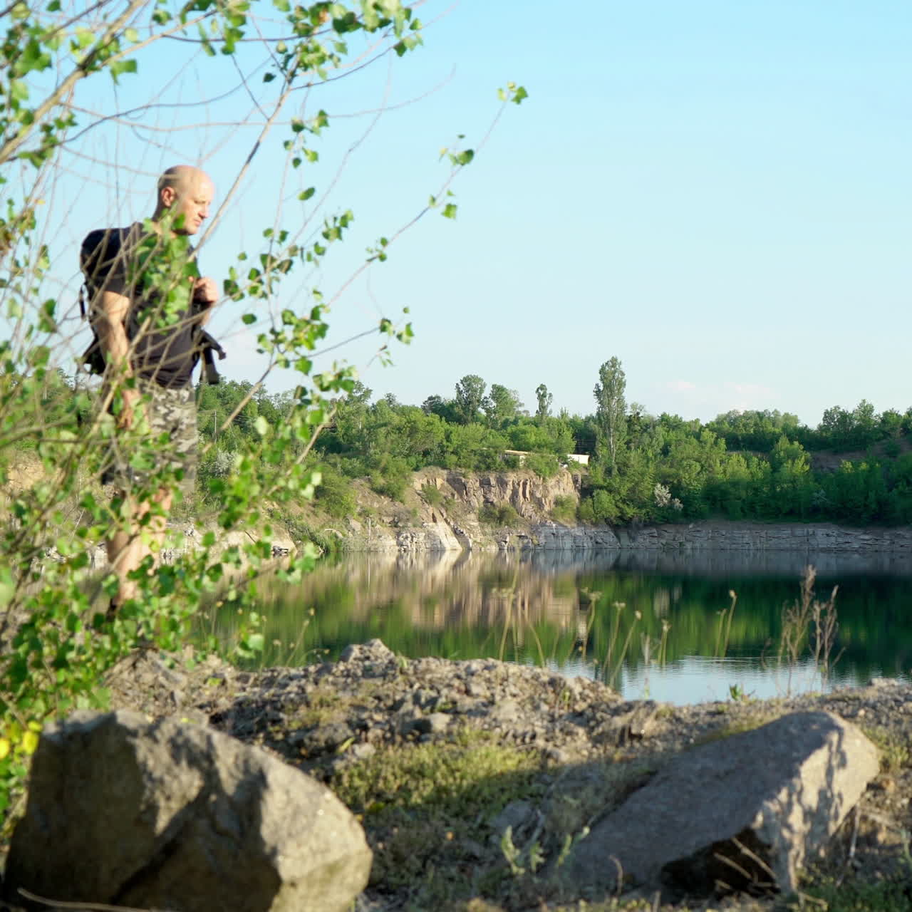 Bald man in t-shirt, shorts and trainers walking with backpack on the big stones near beautiful lake. Natural background of scenic canyon with a clear lake in summer.