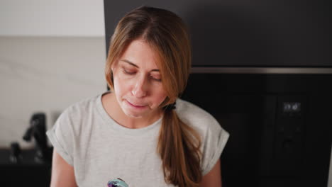 young woman in casual grey t-shirt stands in modern kitchen, concentrating on licking colorful ice cream cone with green waffle cone, standing near fridge and kitchen tap