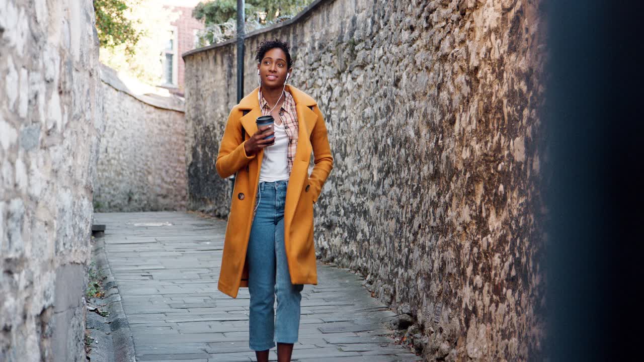 Front view of young black woman wearing a yellow pea coat drinking a takeaway coffee walking in an alleyway between old stone walls, selective focus