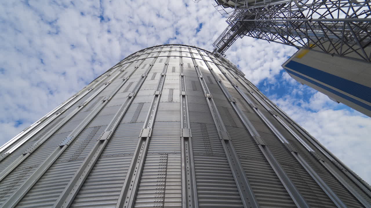 Large metal grain elevator outdoors. Modern silver granary on sky background. Huge aluminum container for storing crop. View from below.