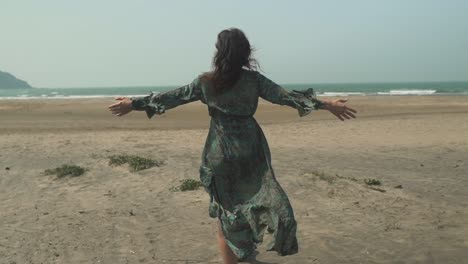 woman on her back walking on the shore of a beach while the wind moves her dress