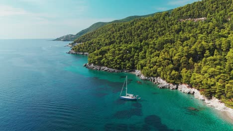 un velero anclado cerca de una costa rocosa y cubierta de pinos en aguas claras de color turquesa bajo un cielo soleado