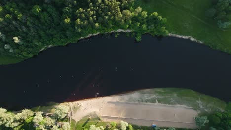 Top-view,-the-river-bed-flowing-between-the-forest-and-the-beach,-people-bathing-in-the-river-on-a-sunny-evening