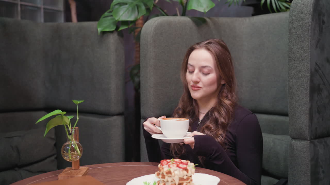 Young woman with long hair gently smiles with closed eyes while holding white cup of latte and saucer close to face, enjoying aroma in quiet indoor setting surrounded by soft green decoration