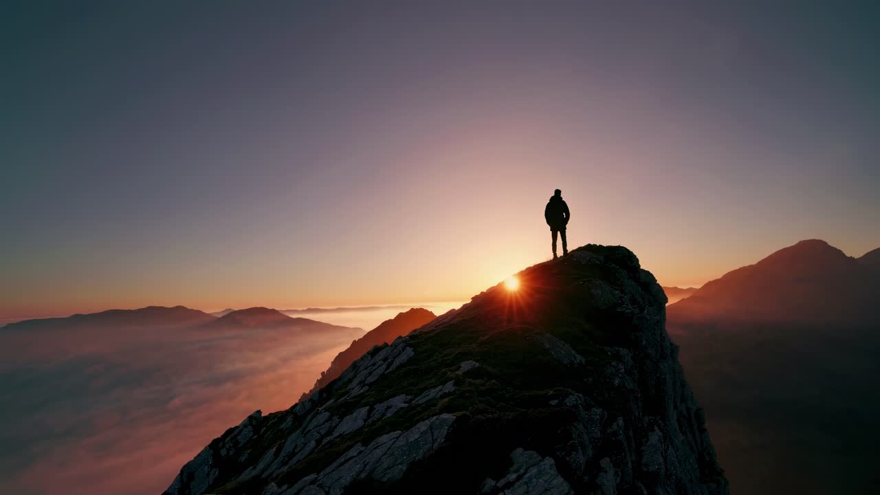 Aerial video captures a lone figure silhouetted against a sunrise atop a mountain peak