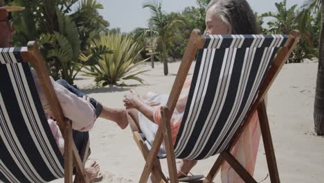 Happy-senior-caucasian-couple-sitting-in-deckchairs-on-sunny-beach-holding-hands,-in-slow-motion