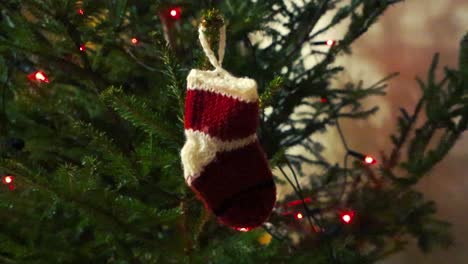 de lana rojo y blanco pequeño calcetín de decoración de navidad colgado en un árbol de navidad verde que ha llevado luz roja en él en el interior. la cámara está girando alrededor de la decoración lentamente manteniendo en de cerca.