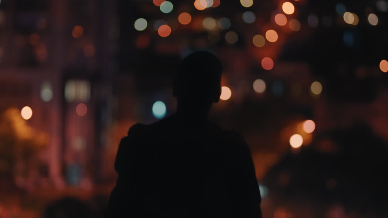 밤에 지붕 위에 팔을 들고 자유를 축하하는 남자의 실루 ⁇  아름다운 도시 불빛을 바라보는 뒷면