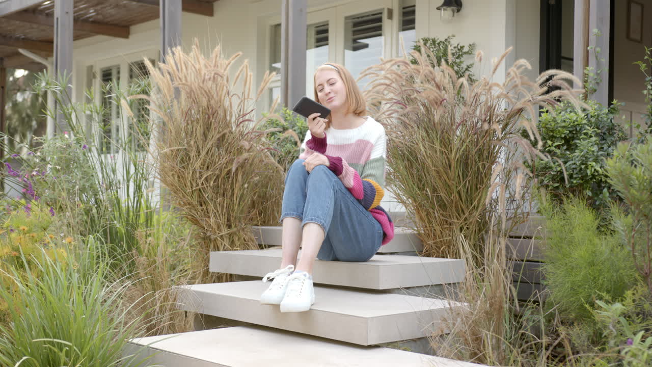 Sitting on steps, woman holding smartphone and smiling in front of house