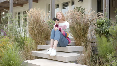 sitting on steps, woman holding smartphone and smiling in front of house