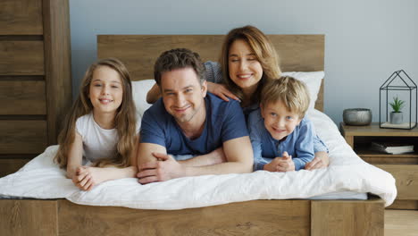 padres alegres y sus hijos acostados en la cama y sonriendo a la cámara por la mañana
