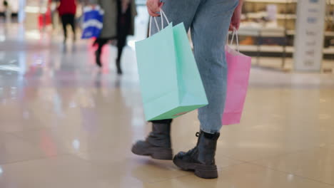 vista lateral de la pierna de una persona con botas negras y vaqueros caminando a través de un centro comercial moderno mientras lleva bolsas de compras, el piso de azulejos elegante y el fondo borroso con otros compradores