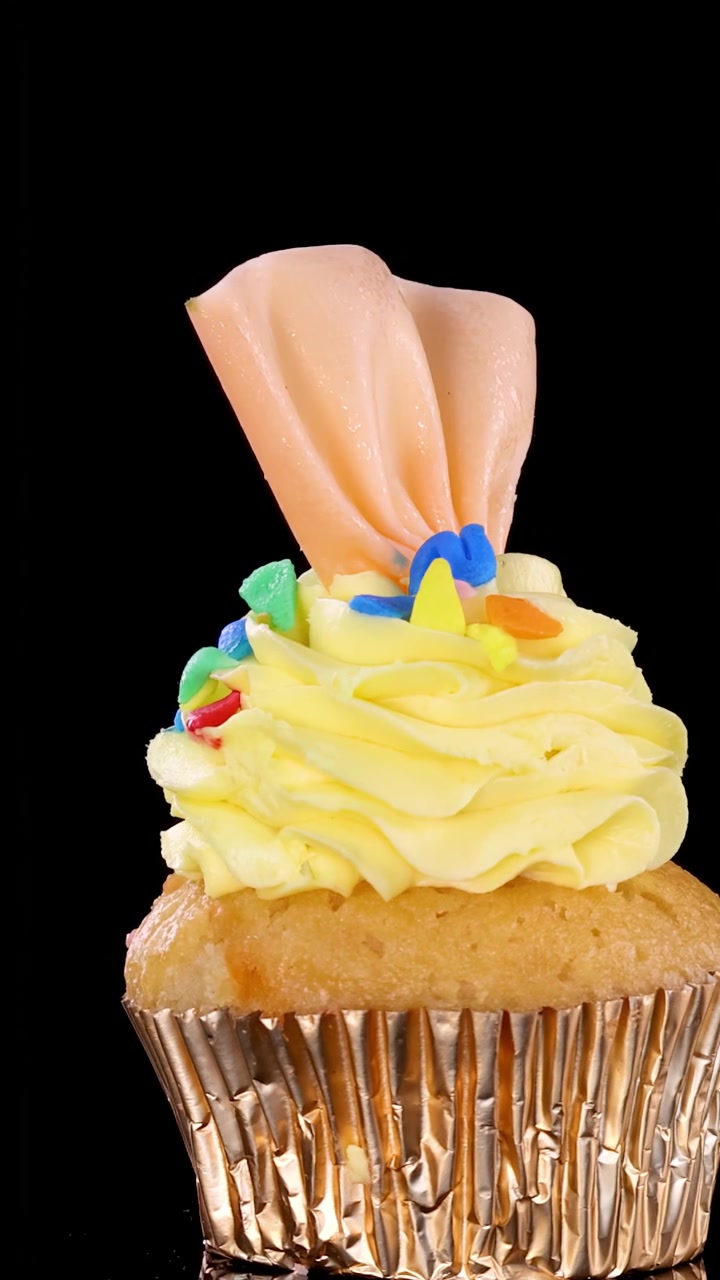 Un singolo cupcake decorato con glassa gialla, zuccherini colorati e una decorazione di caramella rosa ruota dolcemente su uno sfondo nero sotto una luminosa illuminazione da studio.