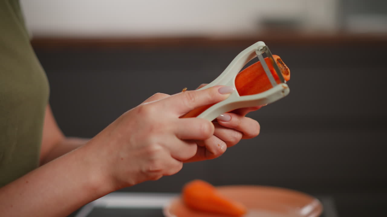 Close up hand of woman in green shirt holding white peeler slicing skin off bright orange carrot over matching plate piled with thin ribbon shavings
