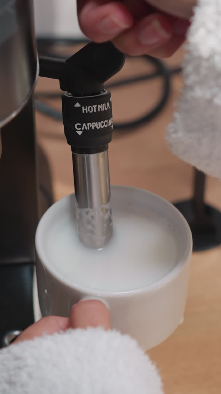 la mujer usa el capuchinador para hacer espuma lechosa. la dama azota la leche en una jarra con un tubo de vapor caliente de la máquina de café en la mesa. proceso de hacer capuchino