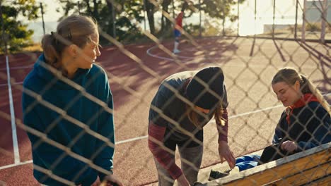 A-happy-trio-of-blonde-girls-basketball-players-in-sports-uniforms-collect-their-things-and-put-them-in-a-backpack-during-their-preparation-for-basketball-practice-on-a-red-summer-street-court-near-a-lattice-fence