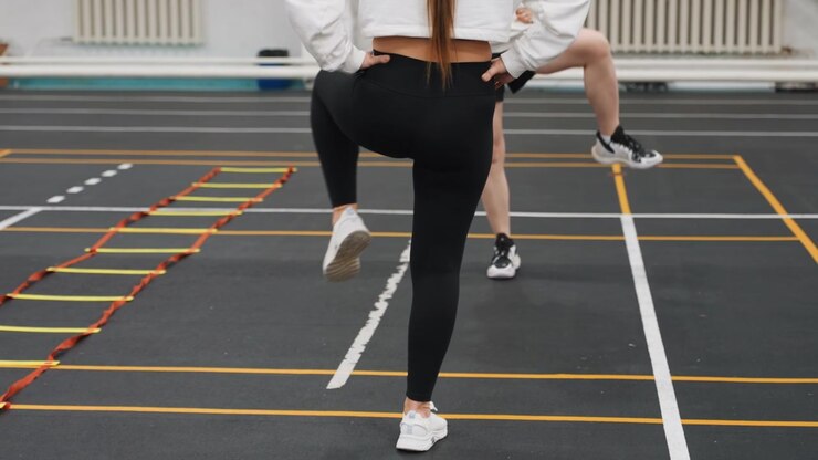 Badminton court agility ladder progression white female athlete rehearsing measured steps and timing across multirung ladder, coach feedback and concentrated repetition on indoor training surface