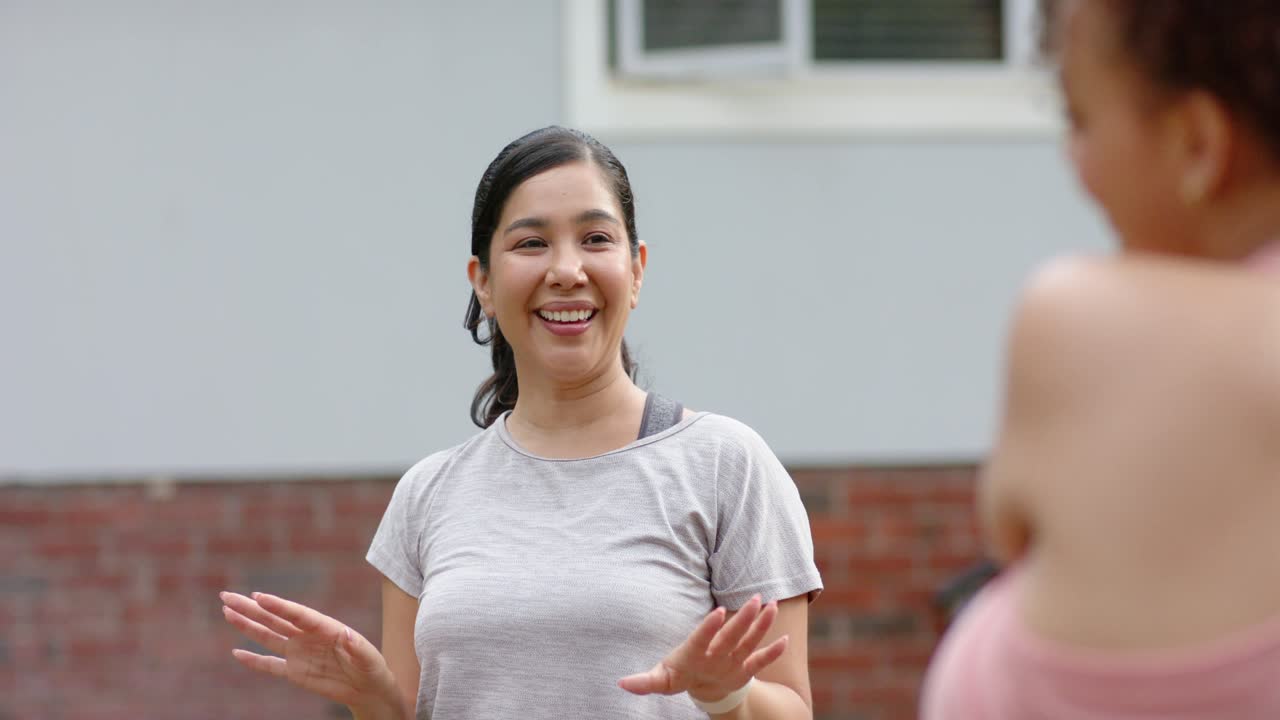 Two women friends talking by house after greeting, gesturing smiling laughing wearing smartwatch