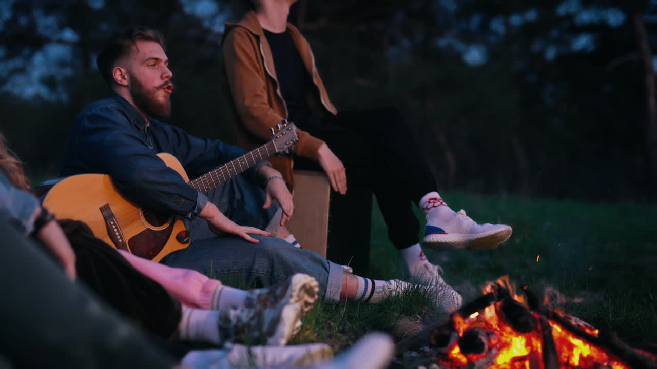 Happy friends spend evening near the campfire. Young people singing and playing the guitar at night. Spending time together in friendly company among nature.