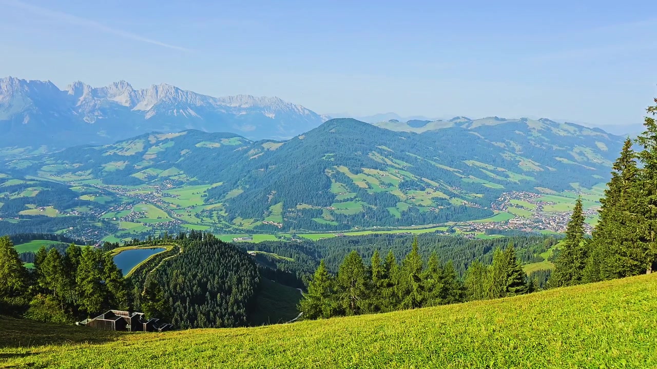 Friedliches Bergpanorama mit saftigen Wiesen, bewaldeten Hängen, entfernten Ortschaften, blauem Stausee und schroffen Alpengipfeln unter klarem Sommerhimmel in einem malerischen Reiseziel in den Alpen.